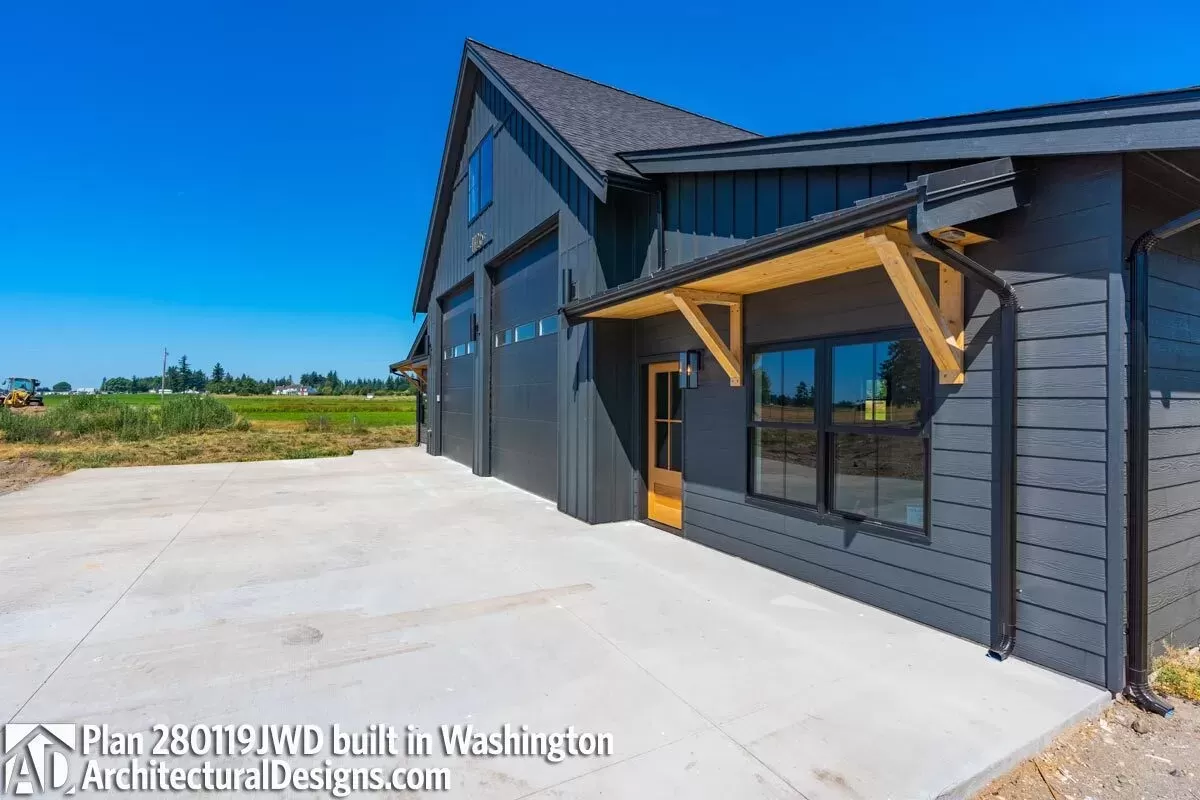 Exterior of a two story garage with a residential loft and ground floor addition with small overhang. Siding is charcoal with natural wooden door and overhand, and a black shingled roof and black modern trimmed windows.