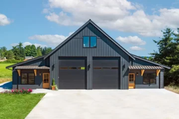Front of a two story garage with a residential loft and two ground floor additions with small overhangs. Siding is charcoal with natural wooden door and overhand, and a black shingled roof and black modern trimmed windows.